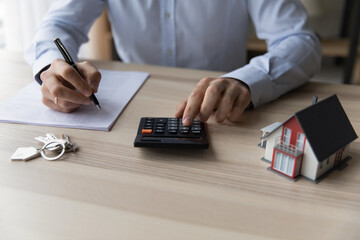 Fototapeta premium Businessman realtor using calculator, sitting at desk with house mockup and keys close up, calculating mortgage rate, preparing documents for contract signing, real estate purchasing concept