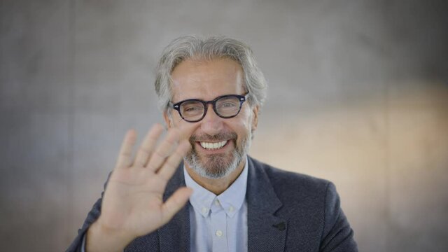 Grey haired man waving to camera. End of video call. Head and shoulders shot. Smiling mature man wearing glasses.
