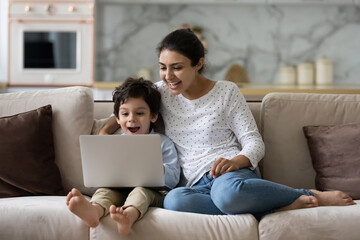 Joyful young indian woman cuddling laughing small cute kid son, watching together funny comedian movie or cartoons online on laptop, relaxing on couch, tech addiction concept.