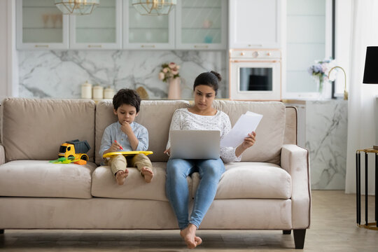 Focused young indian woman working distantly on computer or managing household budget while little kid son playing drawing on toy tablet, sitting together on cozy sofa, parenting and career concept.