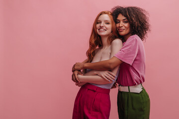 Young african woman warmly hugs her friend from europe with copy space. Cute redhead and brunette are looking at camera cute smiling dressed in bright casual clothes. Home comfort concept - obrazy, fototapety, plakaty