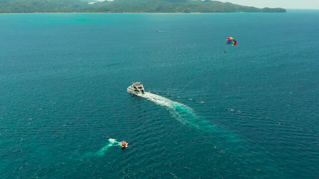 People flying on a colorful parachute towed by a motor boat, aerial view. Parasailing in blue sky.