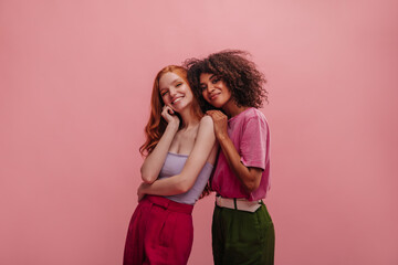 Two interracial young women standing indoors against pink background. Brunette holds red-haired...