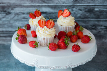 Three cupcakes in the center with white cream decorated with ripe strawberries on a gray blue background and scattered strawberries