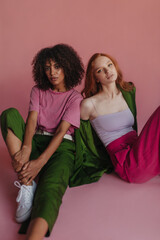 Serious young interracial ladies posing spectacularly while looking at camera against pink background. Brunette and redhead model lean on each other while sitting on floor. Fashionable photo session 
