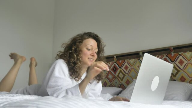 Happy curly haired young woman in white bathrobe falls on bed opens laptop tucking hair behind ears smiles and types waving leg