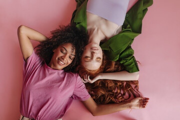 Relaxed young interracial teen girls are lying heads on pink isolated. African lady in T-shirt,...