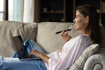 Happy young woman resting on soft cozy couch with gadgets, recording audio message on smartphone,...