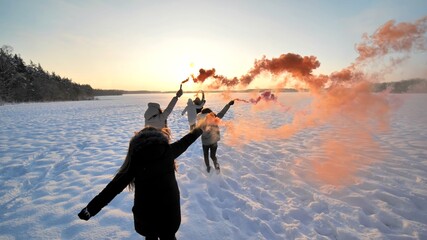 Girls friends run across the winter field with colored smoke.