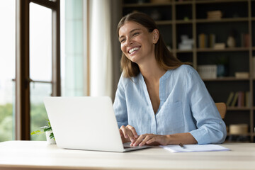 Happy young employee working at laptop, looking away, smiling, laughing. Cheerful woman, female office worker in casual enjoying being at workplace, doing job tasks, talking to colleague, coworker