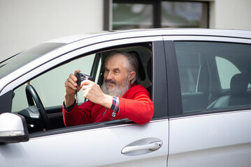 Old man senior with a gray beard is sitting in the car and holding a vintage photo camera