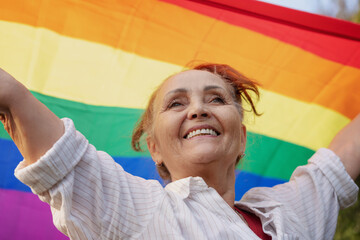 Beautiful mature senior woman with charming smile holding rainbow LGBT flag in her hands, gay and lesbian rights concept - obrazy, fototapety, plakaty