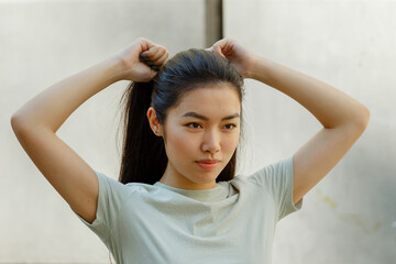 Pretty Asian young woman in t-shirt adjusts ponytail made of long dark hair standing near white wall on sunny day close view - obrazy, fototapety, plakaty