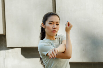 Asian young woman in t-shirt does sports exercises for arms training near light wall with concrete decor on sunny city street