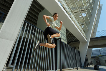 Graceful sportive Asian woman in stylish tracksuit jumping upward moment at training near modern multi-storey parking fence on sunny day