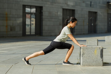 Fototapeta na wymiar Strong Asian woman with long hair in tracksuit does forward dynamic lunges leaning on stone block on city street side view