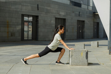 Strong Asian woman with long hair in tracksuit does forward dynamic lunges leaning on stone block on city street side view