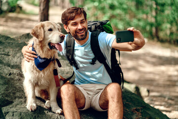 A male hiker with a backpack and a labrador dog makes a selfie on the phone while sitting on a...