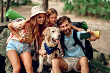Mom and dad with their daughter with backpacks and a labrador dog take a selfie on the phone while...