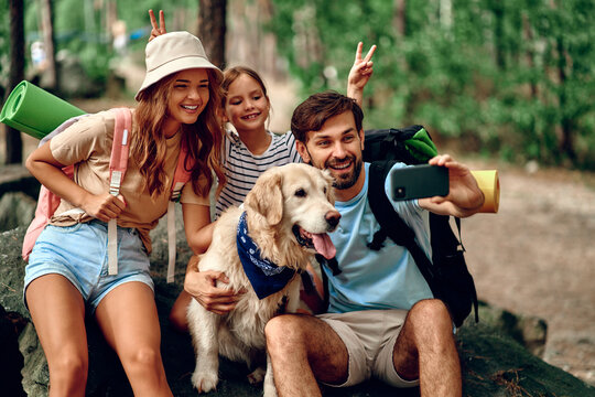 Mom and dad with their daughter with backpacks and a labrador dog take a selfie on the phone while sitting on a stone in the forest. Camping, travel, hiking.
