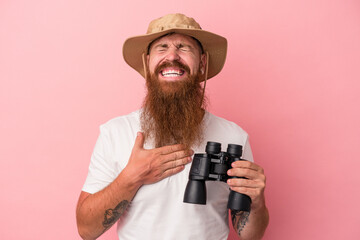 Young caucasian ginger man with long beard holding binoculars isolated on pink background laughs out loudly keeping hand on chest.