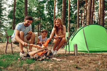 Happy family with a child on a picnic sit by the fire near the tent and grill a barbecue in a pine...