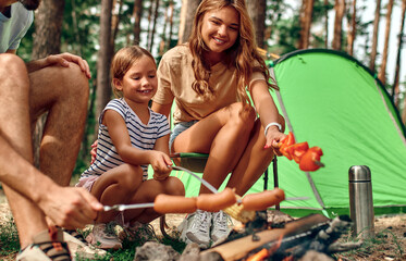 Naklejka na ściany i meble Happy family with a child on a picnic sit by the fire near the tent and grill a barbecue in a pine forest. Camping, recreation, hiking.