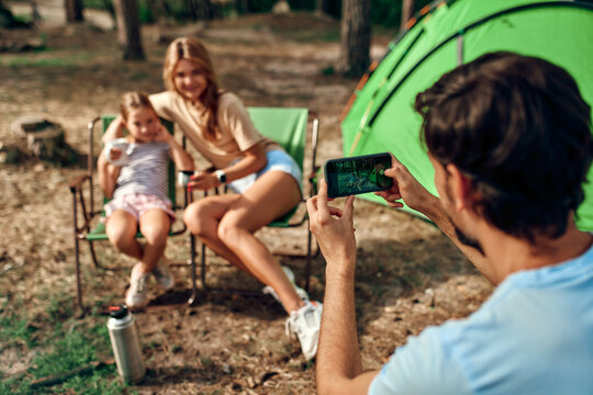 Happy family sitting by the campfire near the tent in the pine forest for the weekend. A man takes a photo of his wife and daughter on his phone. Camping, recreation, hiking.