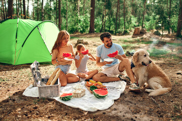 Happy family with a child and a dog at a picnic sit on a blanket near the tent and eat fried food...