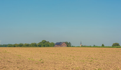 Fototapeta premium Bare farmland with a house, a church and trees on the horizon under a clear blue sky.