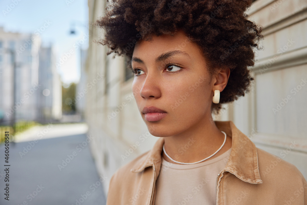 Wall mural Close up shot of good looking thoughtful woman with Afro hair focused into distance with serious expression spends free time outside thinks about future plans. People leisure lifestyle concept - Wall murals