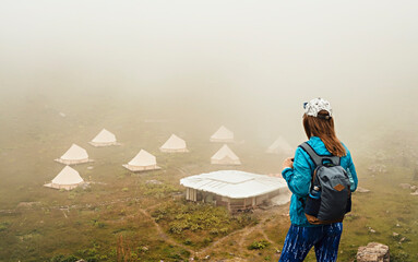 Obraz premium rear view of young woman in cap and blue sportswear with backpack hiker standing in green mountain valley and looking at tent camping in fog, hiking