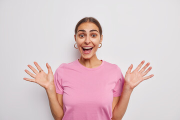 Happy cheerful woman raises palms feels glad smiles cheerfully reacts on amazing news dressed in casual pink t shirt isolated over white background. People human reactions and feelings concept