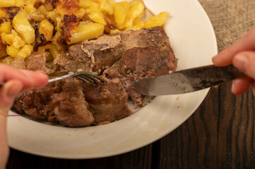 Someone is cutting a piece of juicy roast meat on a plate of fried potatoes with a knife and fork. Close-up Selective focus.