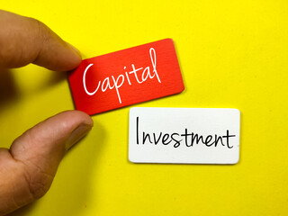 Hand holding colored wooden board written with text Capital Investment on a yellow background.