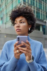 Pensive dark skinned woman blogger uses media application on smartphone gadget enjoys journey trip for visiting city dressed in casual blue jumper poses in urban setting has online communication