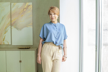 Cute smiling senior woman in blue casual blouse standing near window