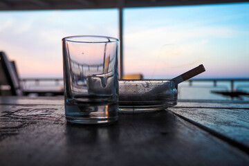 Consequences of the party. Empty beer glass and smoking cigarette in dirty ashtray at sunrise