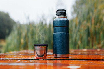 Thermos and mug with hot drink standing on wet wooden table after rain outdoors, close-up. Steam rises from thermo mug. Warming drink from hiking flask in cold, rainy weather