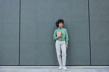 Fototapeta na wymiar Full length shot of pensive Afro America millennial girl dressed in fashionable clothes looks away listens music in headphones holds diary poses against grey wall with blank space for your advert