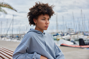 Photo of beautful thoughtful young Afro American woman dressed in hoodie poses on bench at pier...