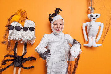 Little children and halloween concept. Little child wrapped with white fabric black spider web tries to looks spooky frightens people poses against decorated orange background. 31st October is coming