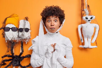 Surprised young woman dressed like mummy or ghost celebrates halloween afraids of something scarying poses against orange background creepy creatures near. Ethnic female in carnival costume.