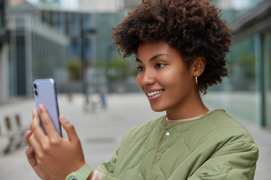 Horizontal shot of happy Afro American woman makes video call via smartphone connected to high speed internet wears jacket enjoys good weather poses against blurred background. Lifestyle concept