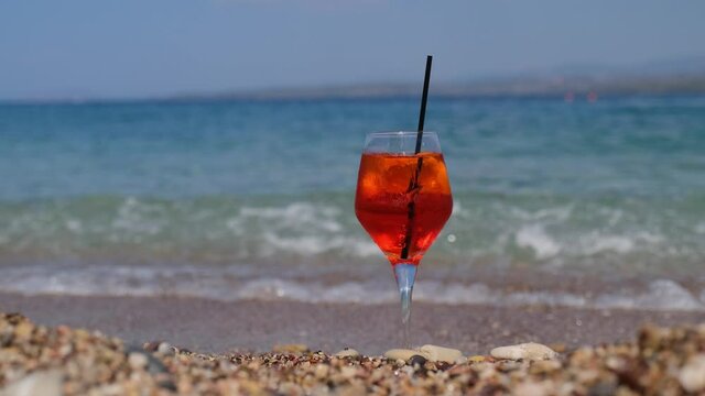 glass of aperol spritz cocktail stands on sand near the sea