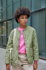 Serious beautiful curly haired woman wears stylish jacket trouses poses outside with pensive expression looks away discovers new places of interest during excursion time. Recreation concept.