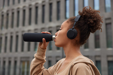 Sideways shot of curly haired Afro American woman drinks water during workout break dressed inn sportsclothes listens audio track in wireless headphones poses outside against building background