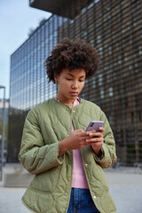 Fototapeta na wymiar Vertical shot of serious Afro American woman uses mobile phone for sending messages searches web publication connected to wireless internet dressed casually poses in city centre uses chat application