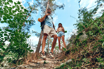 Naklejka na ściany i meble A loving couple of tourists with backpacks climb the rocky peaks in the forest. Hiking, traveling, hiking.