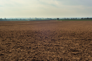 Bare arable field with a skyline of trees and a city under a cloudy sky.